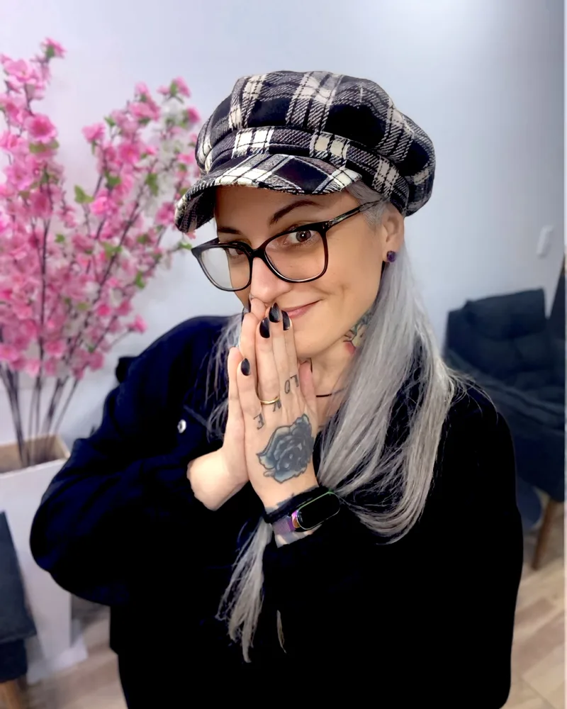 Joana Chung de boina xadrez e óculos, com as mãos unidas em frente ao rosto, mostrando a tatuagem de rosa na mão